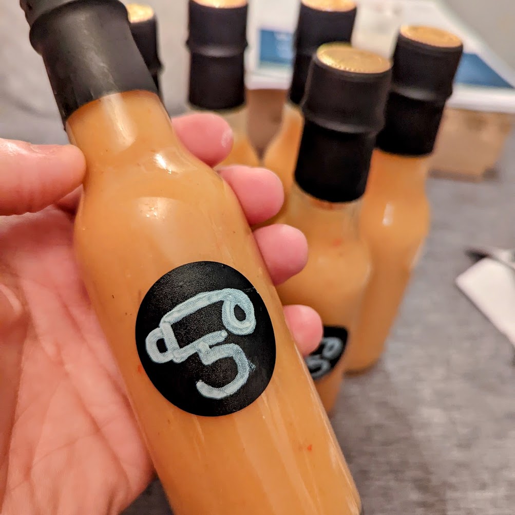 Holding a bottle of hot sauce with several more in the background. The color of the sauce is nearly brown with label that has a rough character drawing of a face.