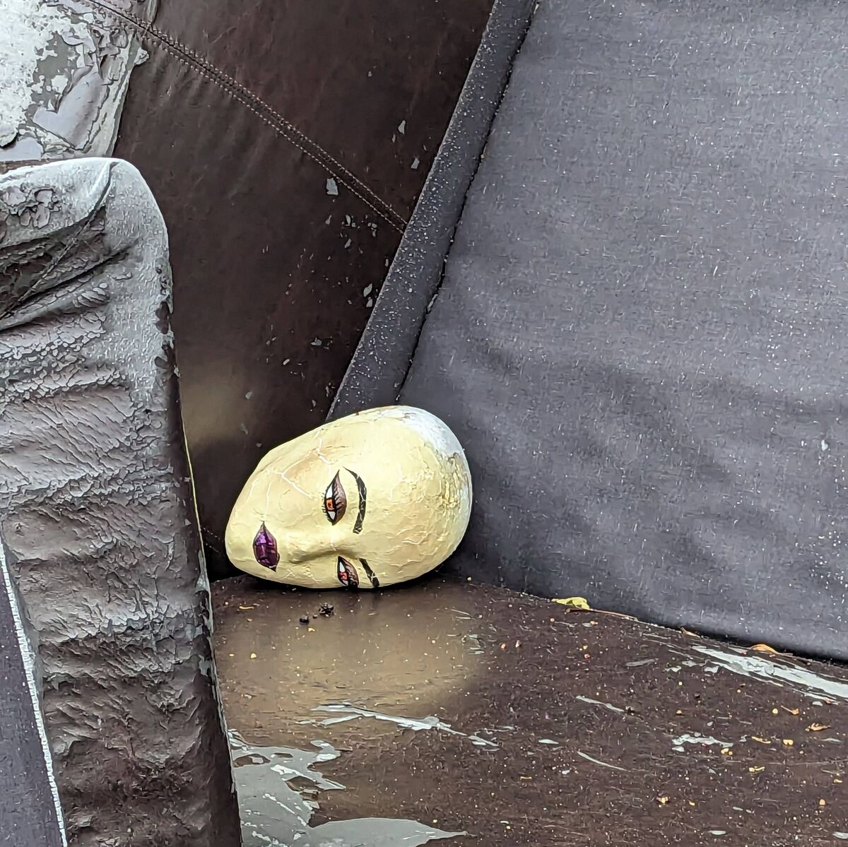 A mannequin head sitting on a couch by the trash - looks like it was used for practicing doing makeup on.