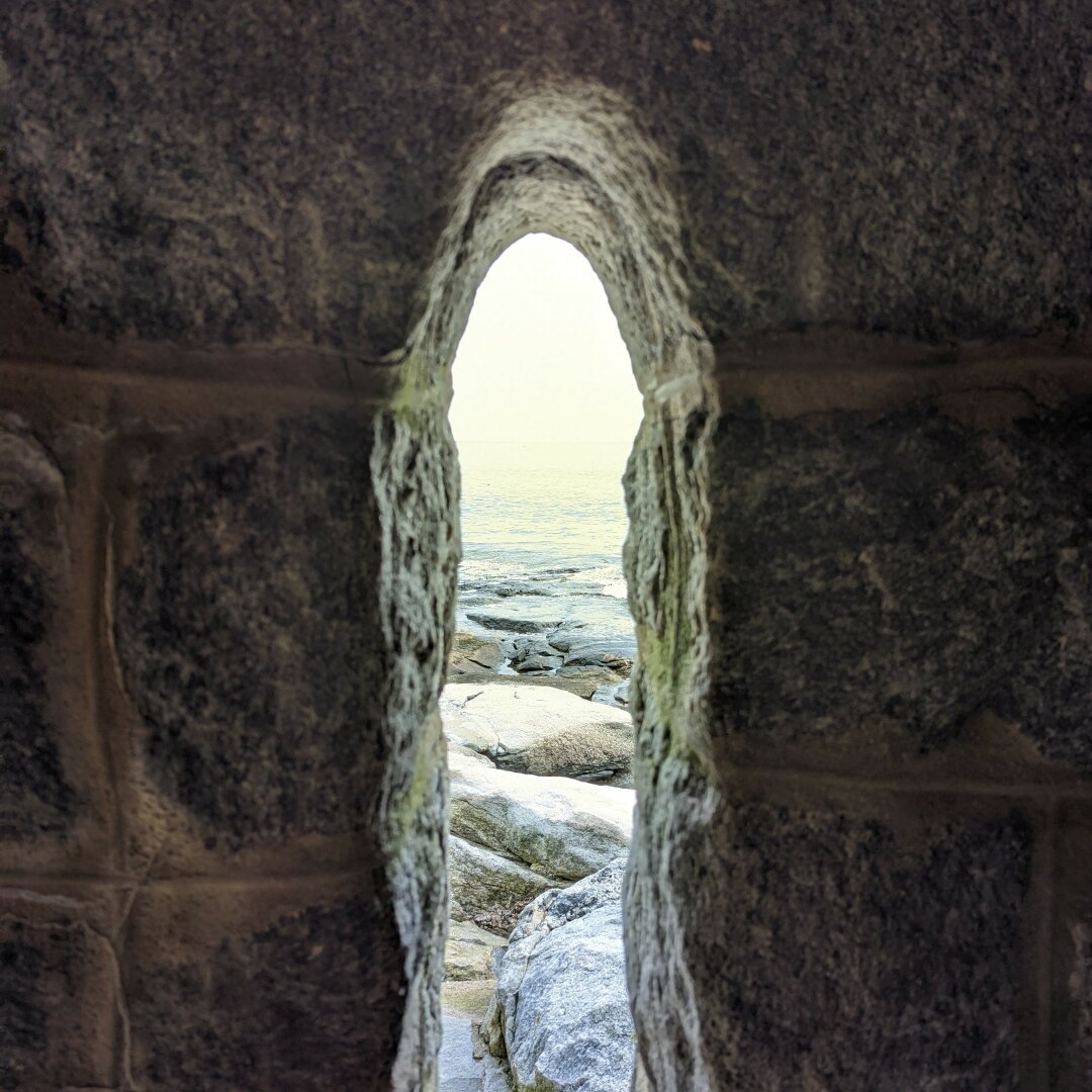 Inside a stone building looking out of a small thin window over some large rocks at the edge of the ocean