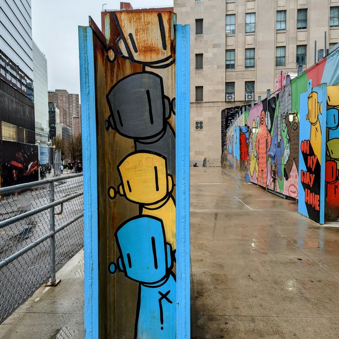 Graffiti of 4 characters that look like robots painted on a metal beam standing upright in them middle of the sidewalk