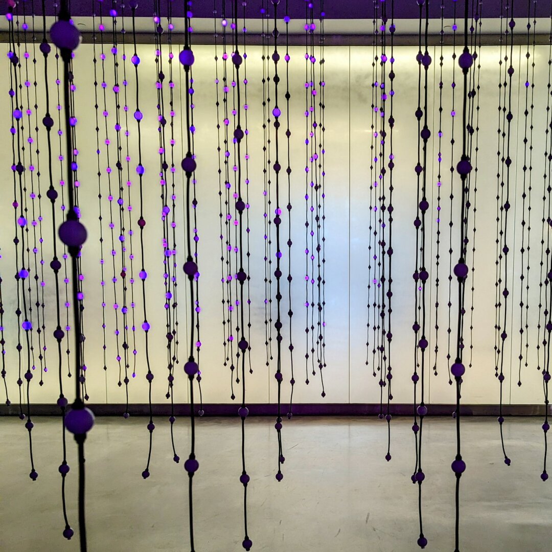 A muted cream colored room with hundreds of led lights hanging in the foreground. Most are off, but others are glowing purple