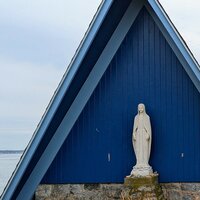 A chapel with a very steep blue steeple with a statue of a women in front of it, the ocean off in the background