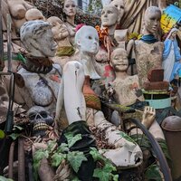 a Collection of decayed and weather damaged mannequins sitting upright together in a garden against a chainlink fence