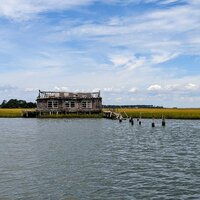 An abandoned house in the middle of marshland sitting up against the water - the roof is collapsed and their is remnants of a dock