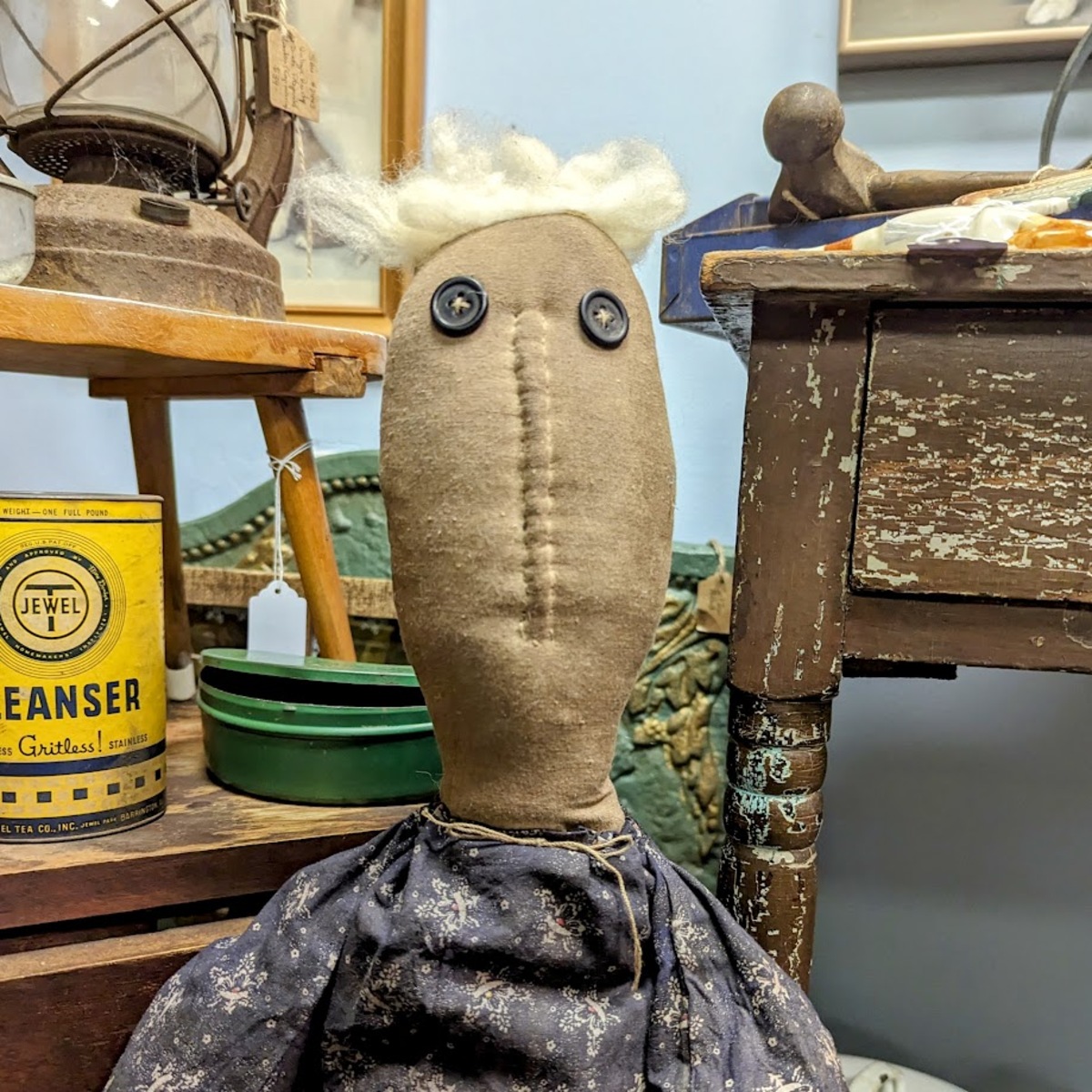 A doll, sitting among old coffee cans and worn down furniture, with a long face, blonde tufts of hair, button eyes and a vertical mouth stretching from chin to forehead. 